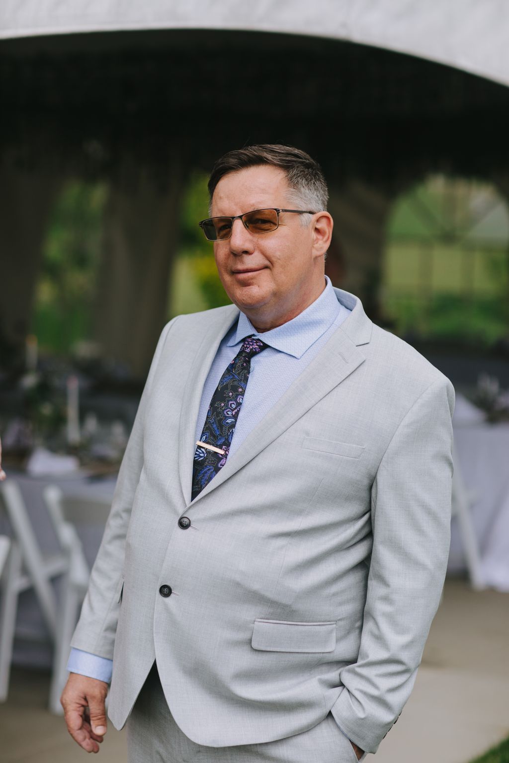 Groom in light gray blazer and patterned tie wearing sunglasses, posed outdoors with blurred green foliage background