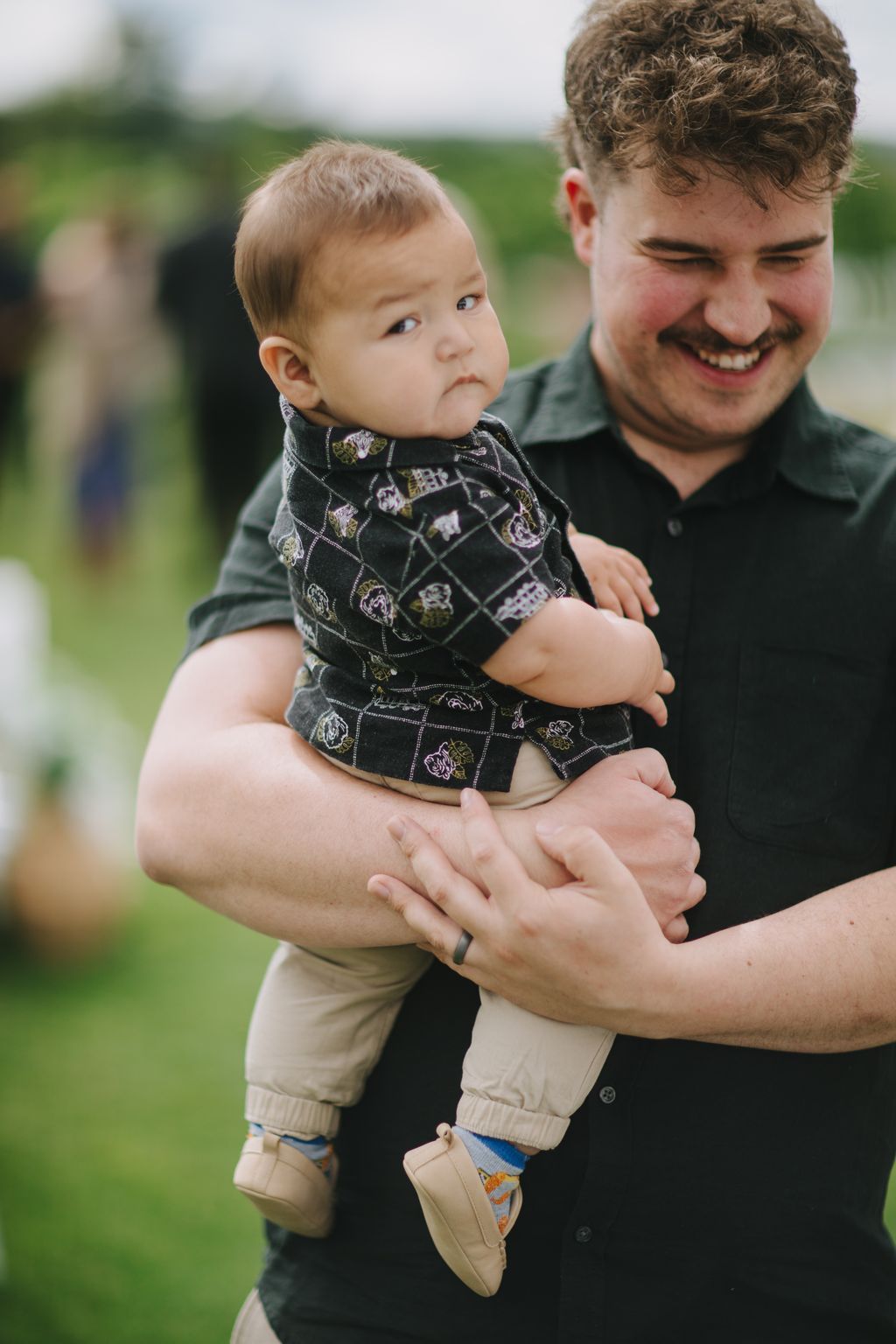 Father holding baby son in patterned shirt outdoors with green field background