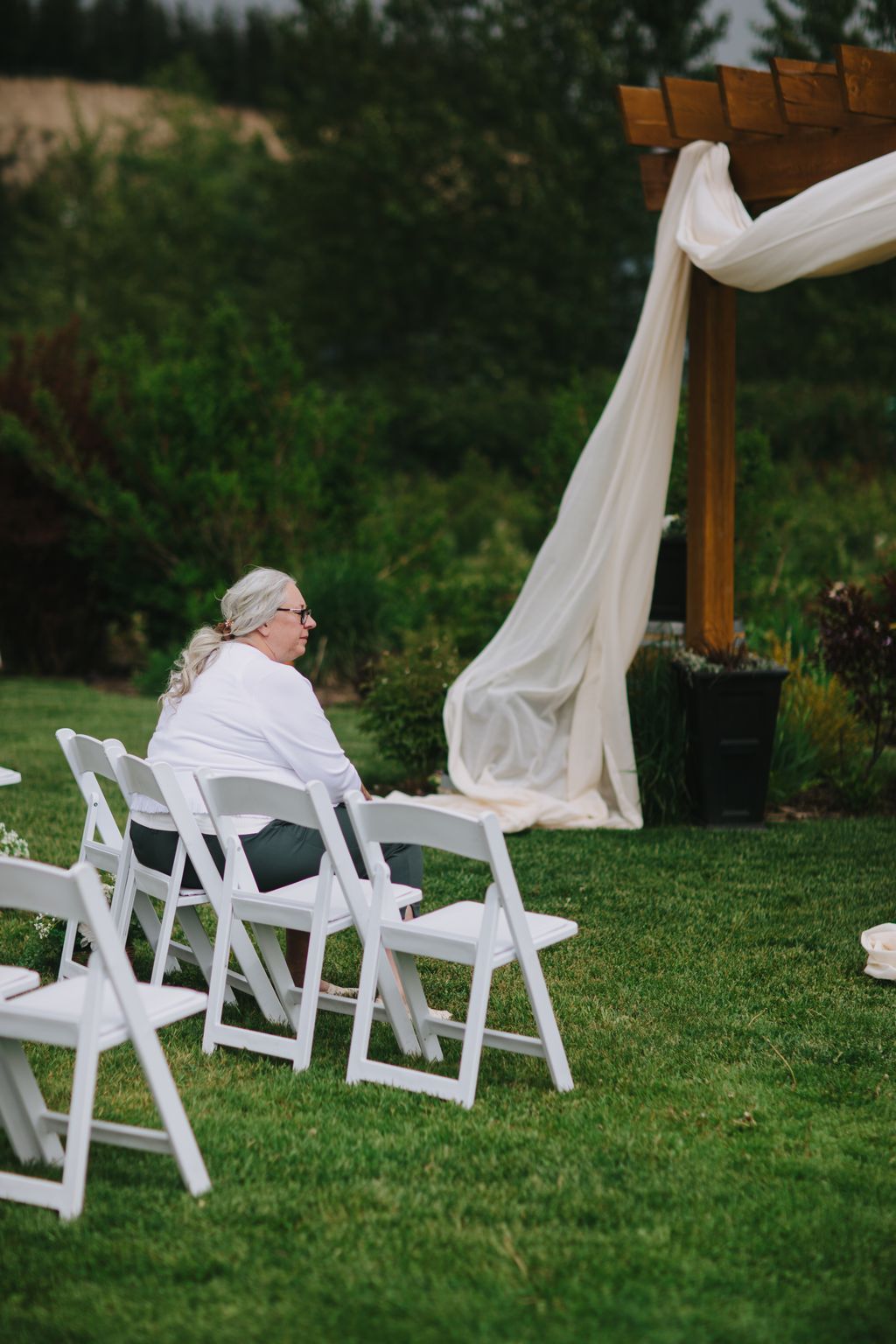 Elderly woman in white shirt seated on white folding chair on manicured lawn, gazing at flowing cream fabric draping from wooden structure with evergreen tre…