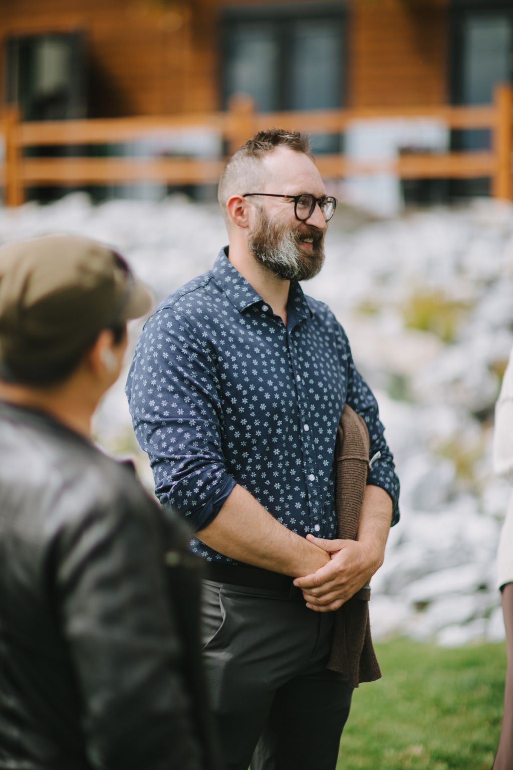 Groom in navy snowflake-patterned shirt and glasses smiles while standing outdoors near a brick building