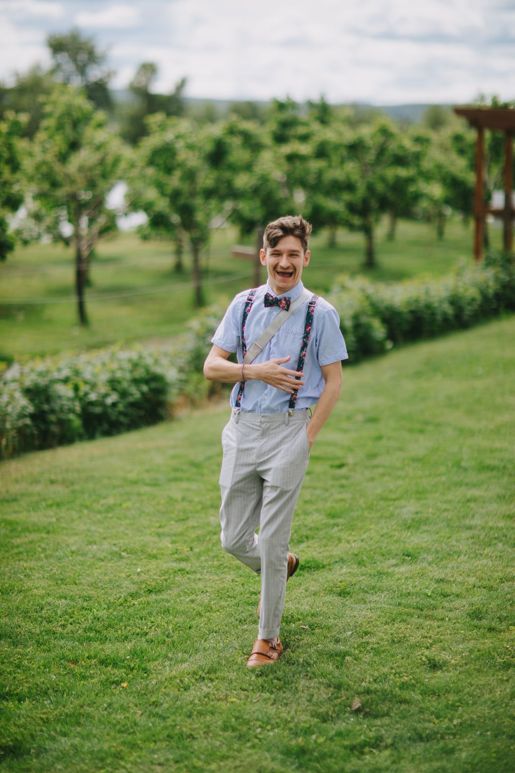 Groom in light blue shirt, suspenders and bow tie running through orchard grass with trees in soft focus background