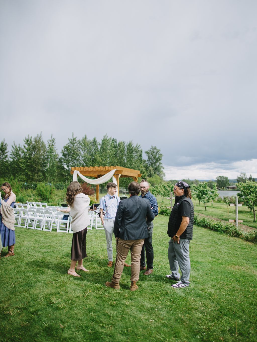 Guests gather under a wooden pergola with white draping on an overcast day at an outdoor garden wedding ceremony