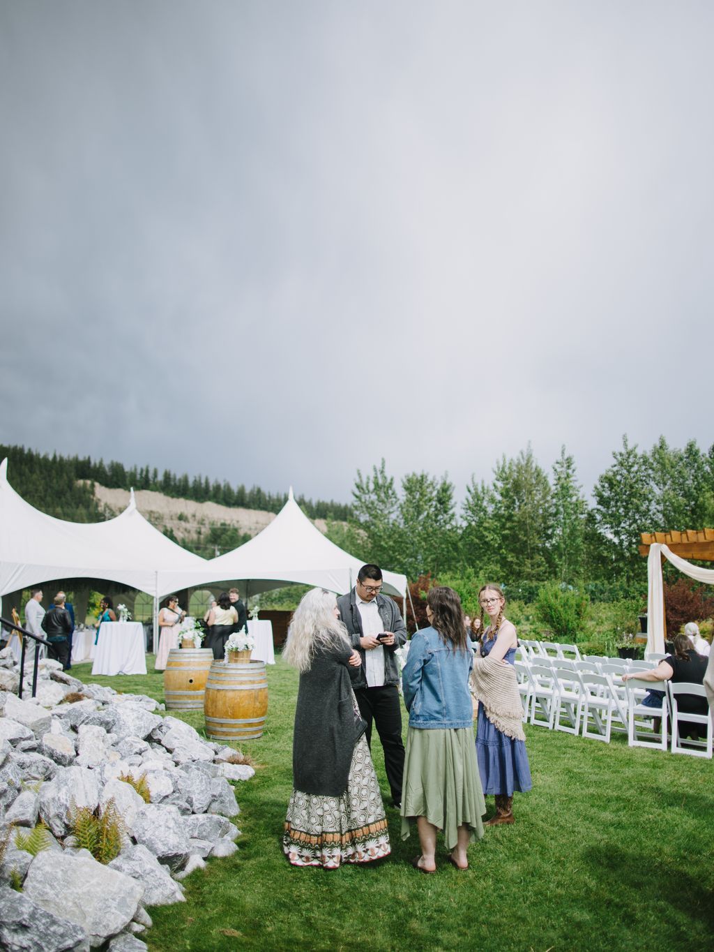 Guests gathered on a grassy lawn beneath a white tent with forested mountains in the background during an outdoor wedding reception