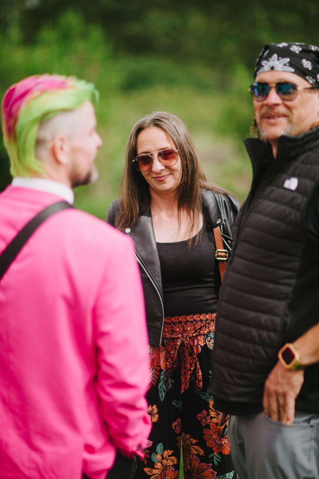 Woman in sunglasses and black leather jacket smiles at companion in pink, green blurred background