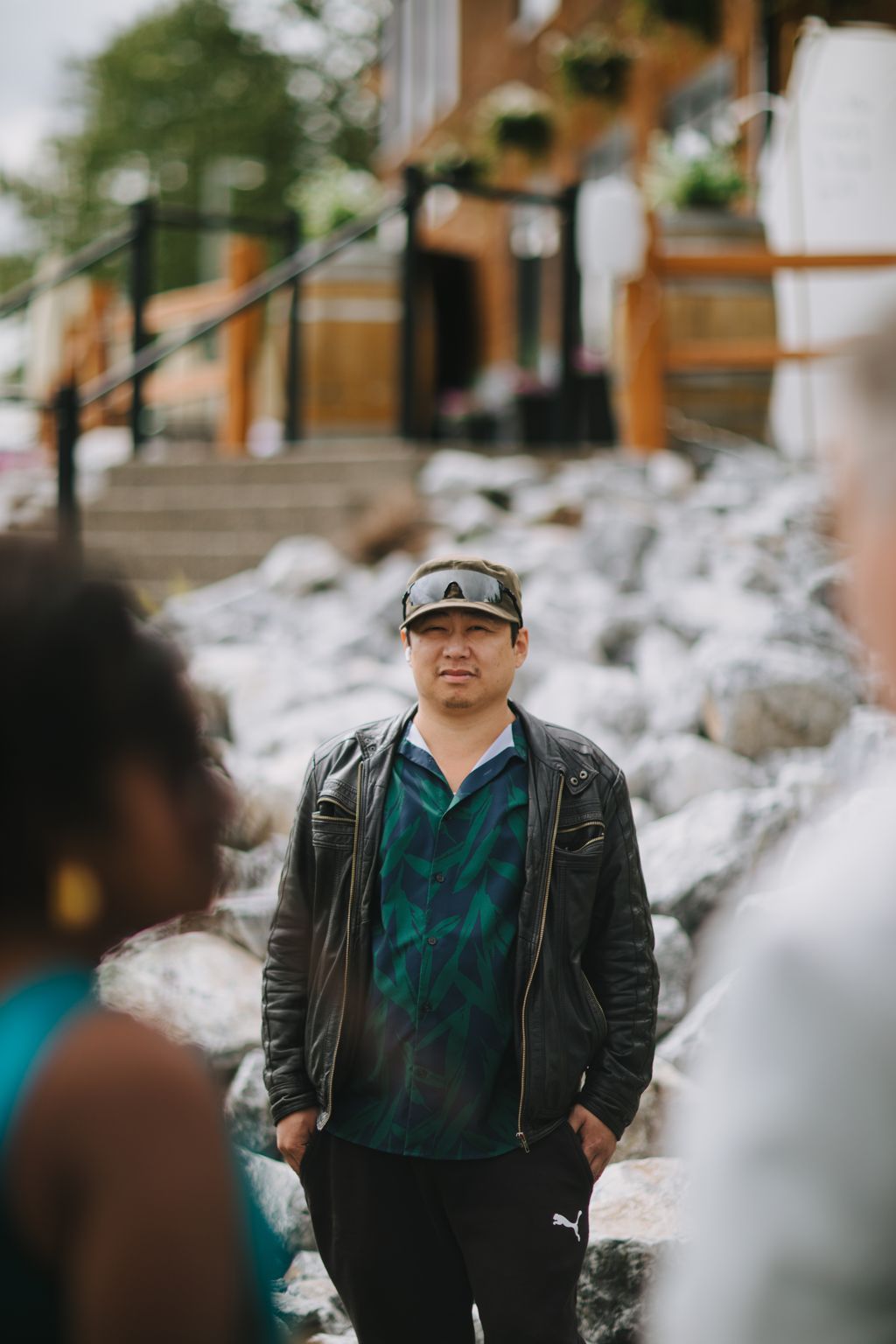 Man in black leather jacket and cap standing on stone steps with blurred figures and warm-toned building in background