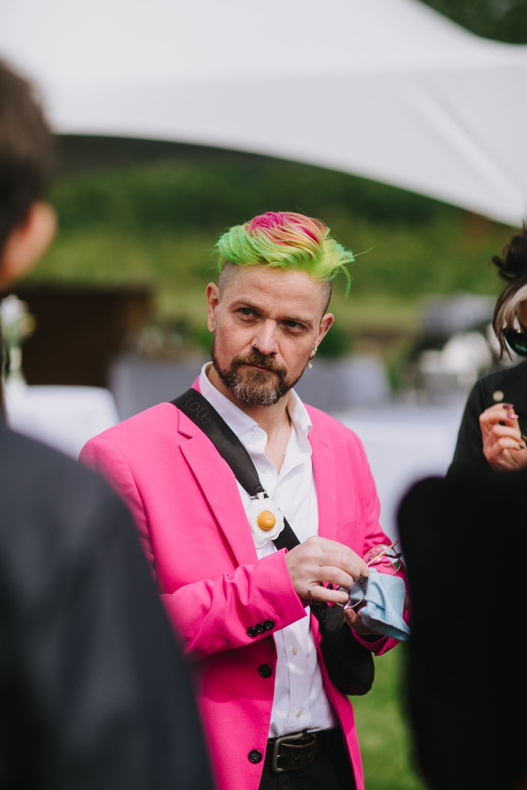 Groom in vibrant pink blazer and neon green hair poses outdoors with soft-focus green landscape backdrop