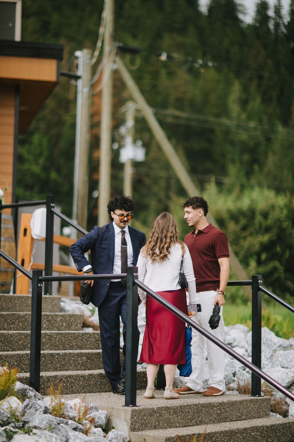 Three guests converse on outdoor stairs beside a metal railing, surrounded by lush green forest in soft natural light