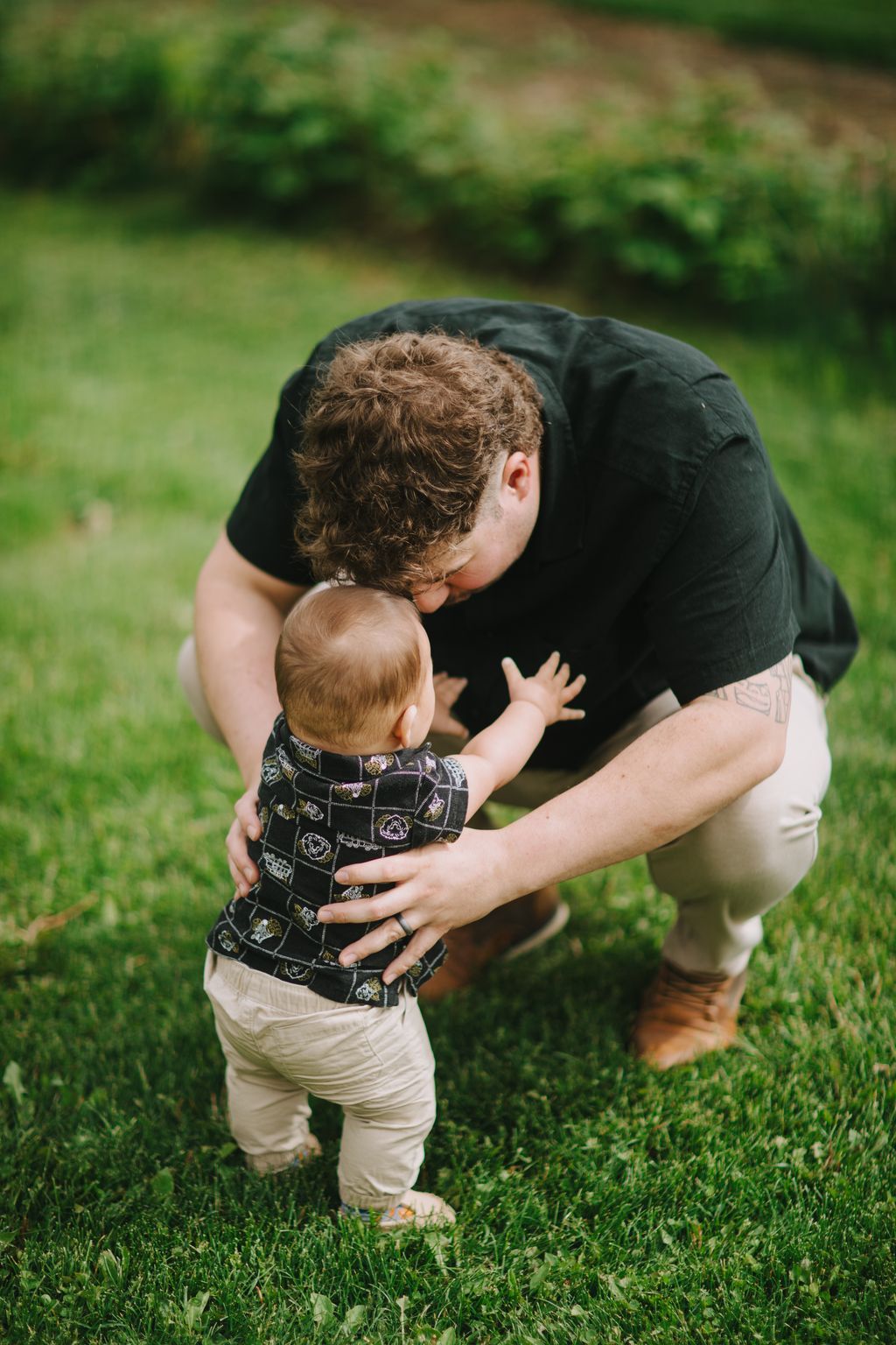 Older brother holds toddler on sunny grass, both with curly brown hair, intimate family moment outdoors