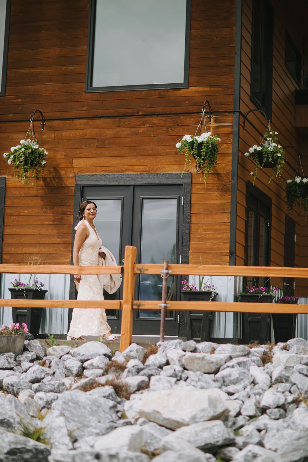 Bride in ivory dress stands by modern wooden cabin with hanging white flowers and stone pathway details