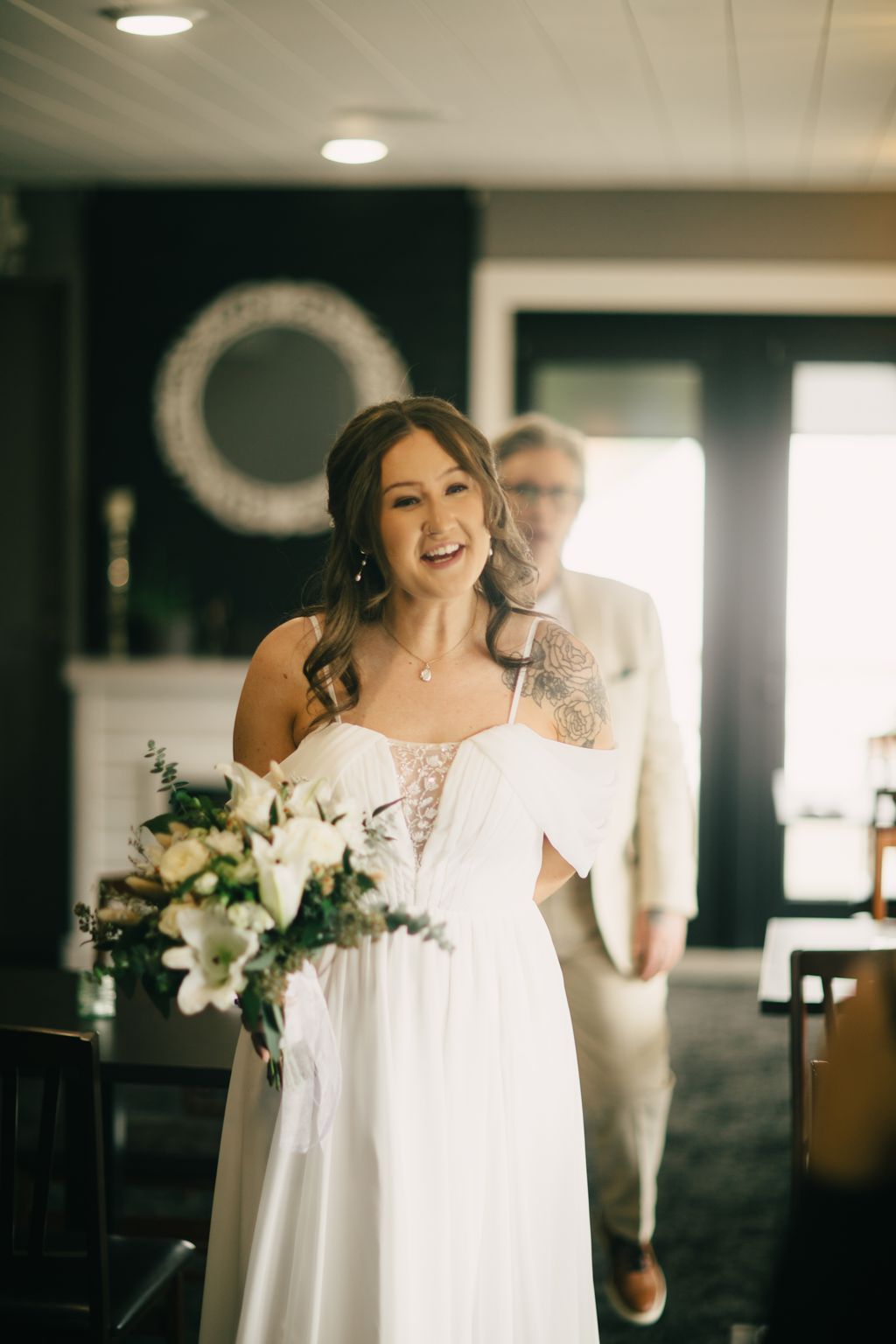 Bride in flowing white off-shoulder dress with lace detail smiles while holding bouquet in modern indoor venue with recessed lighting