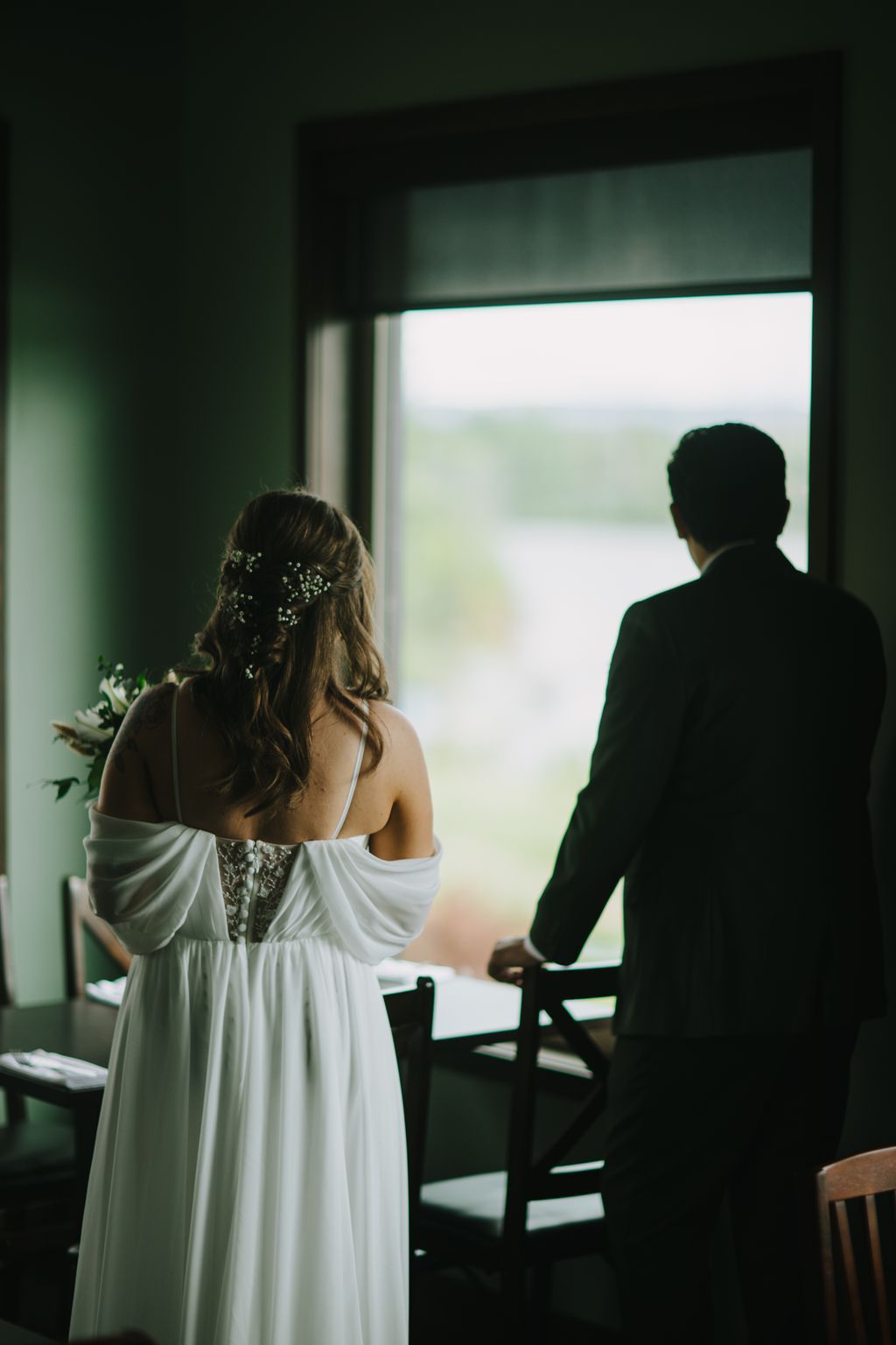 Bride in white dress with baby''s breath hair clip stands beside groom in dark suit, backlit by bright window with soft natural light