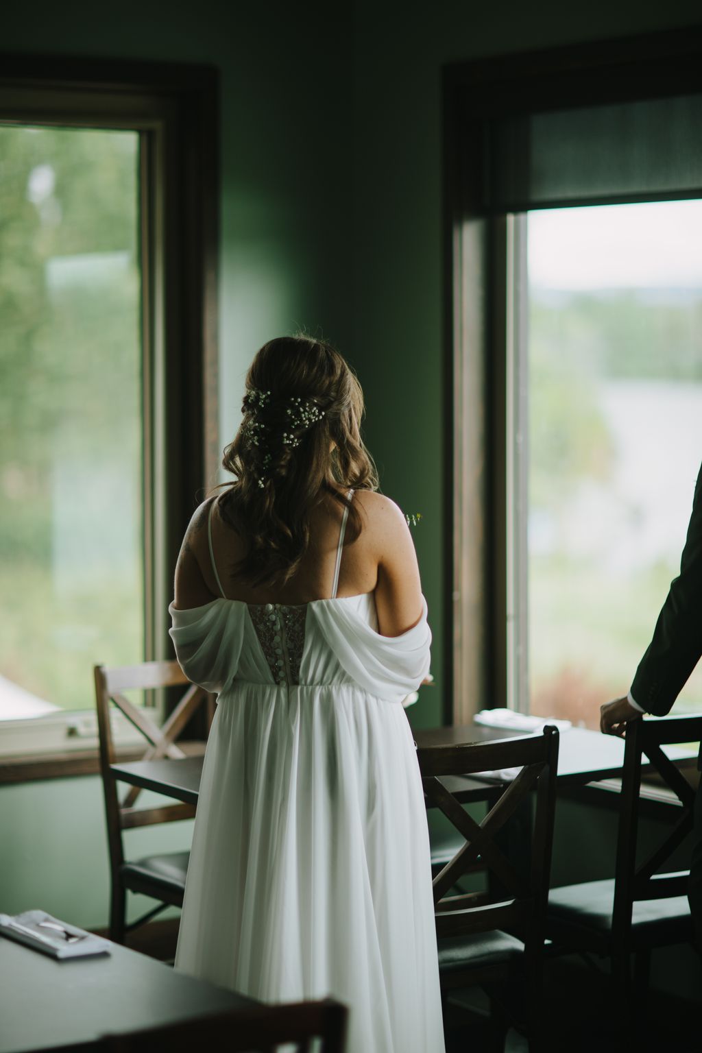 Bride in flowing white dress with baby''s breath in her hair, viewed from behind, standing in soft natural light before a green wall