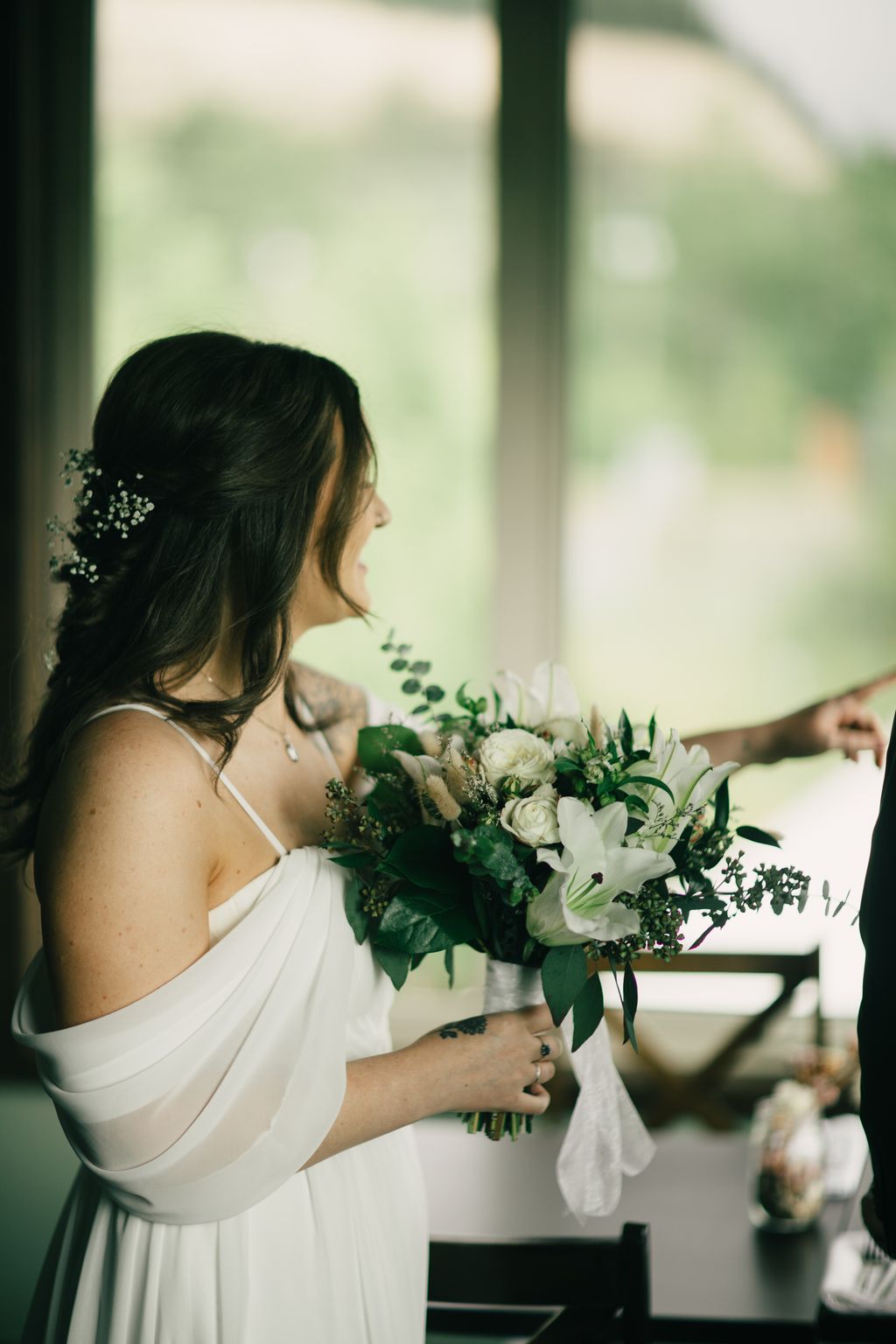 Bride in white dress holding bouquet of white roses and greenery, photographed in profile near a bright window with soft natural light