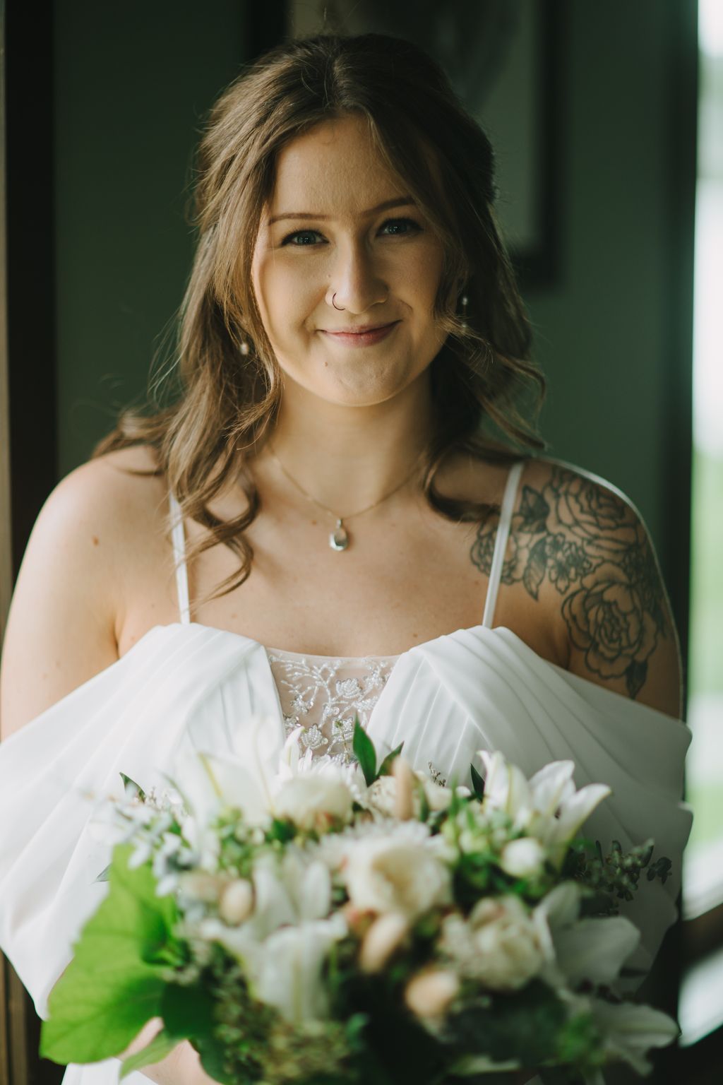 Bride in white lace off-shoulder dress holding white bouquet, smiling warmly in soft natural light