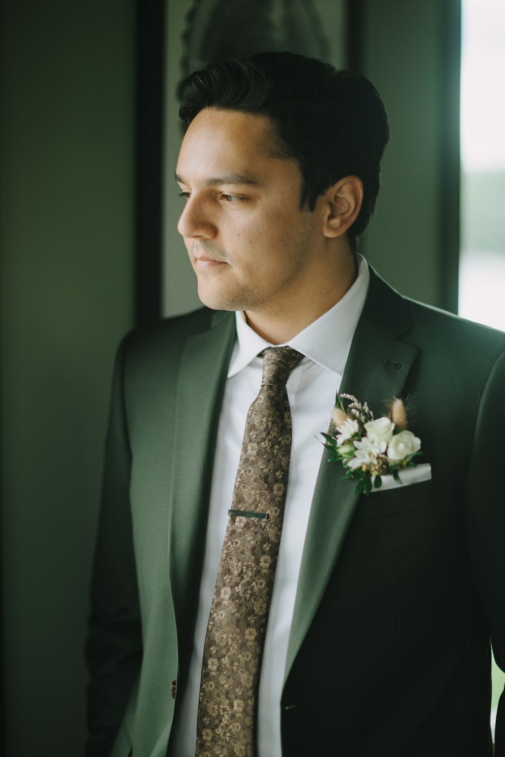 Groom in sage green suit and patterned tie with boutonniere, gazing thoughtfully to side against soft green background