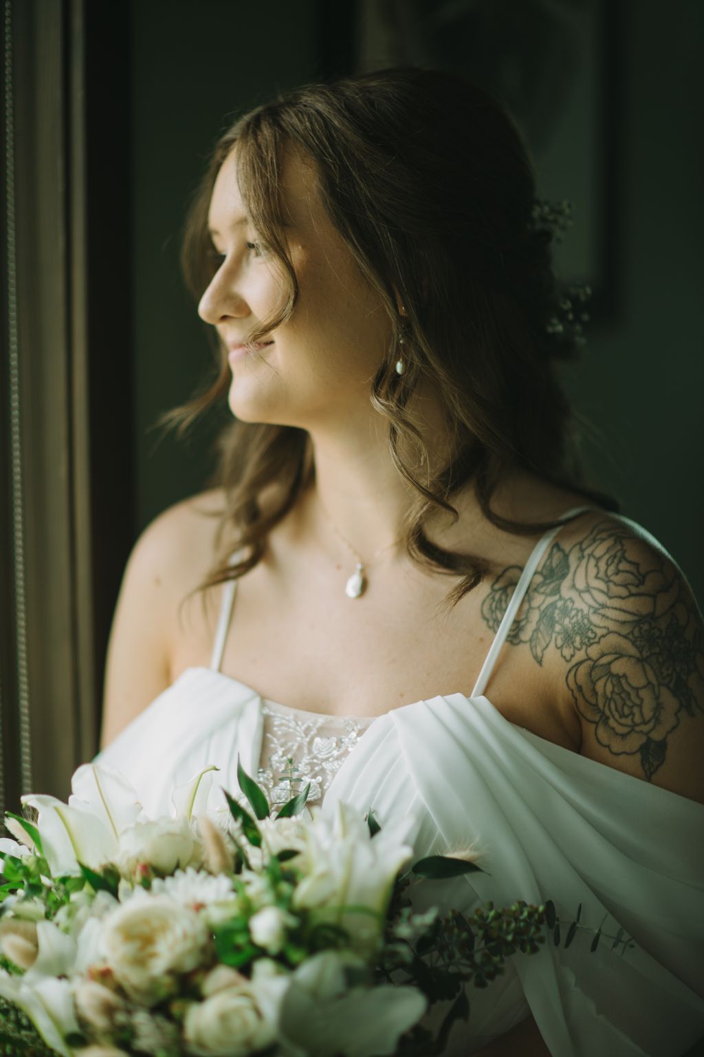 Bride in white off-shoulder gown with pearl jewelry gazes downward, holding white flowers, warm golden light against dark background