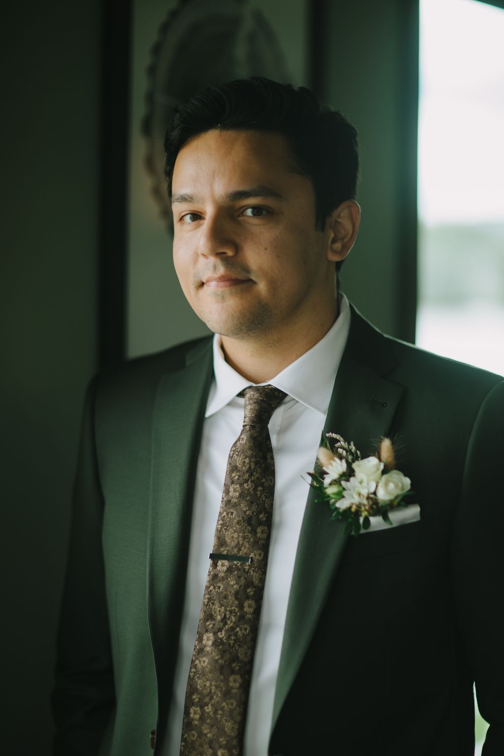 Groom in sage green suit and patterned tie with boutonnière against green backdrop, looking thoughtfully to the side