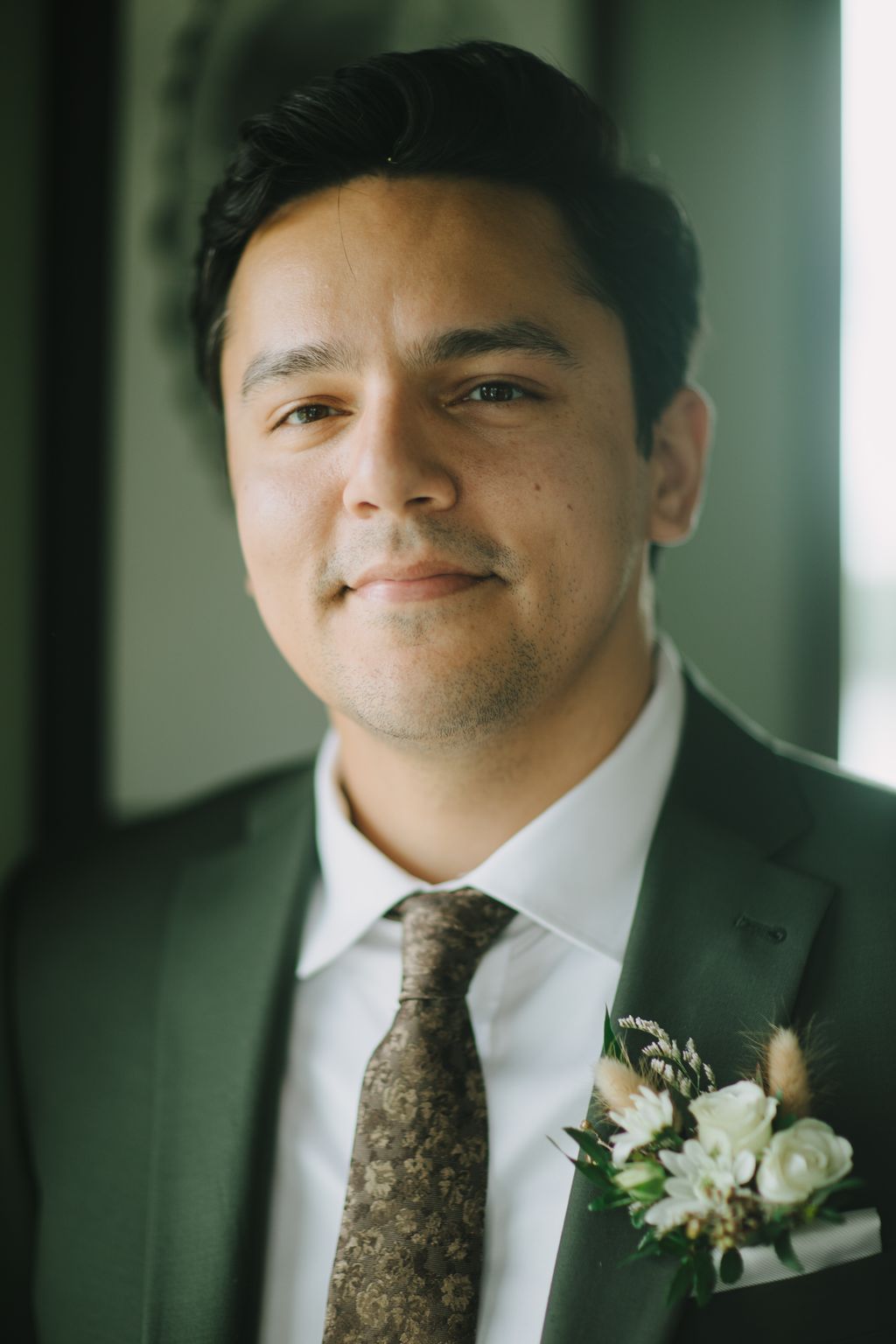 Groom in sage green suit and patterned tie with boutonniere, soft-focused green background, warm natural light