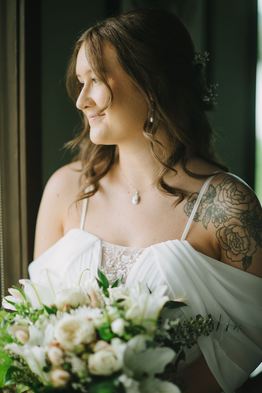 Bride in white off-shoulder dress with pearl necklace holds white bouquet, gazing thoughtfully in soft natural light