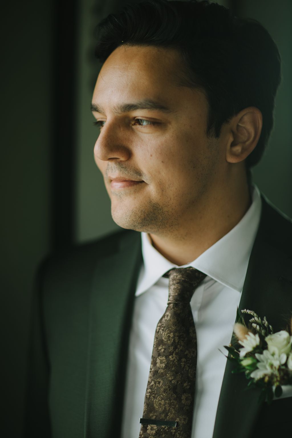 Groom in profile wearing white dress shirt and patterned olive tie against soft green background