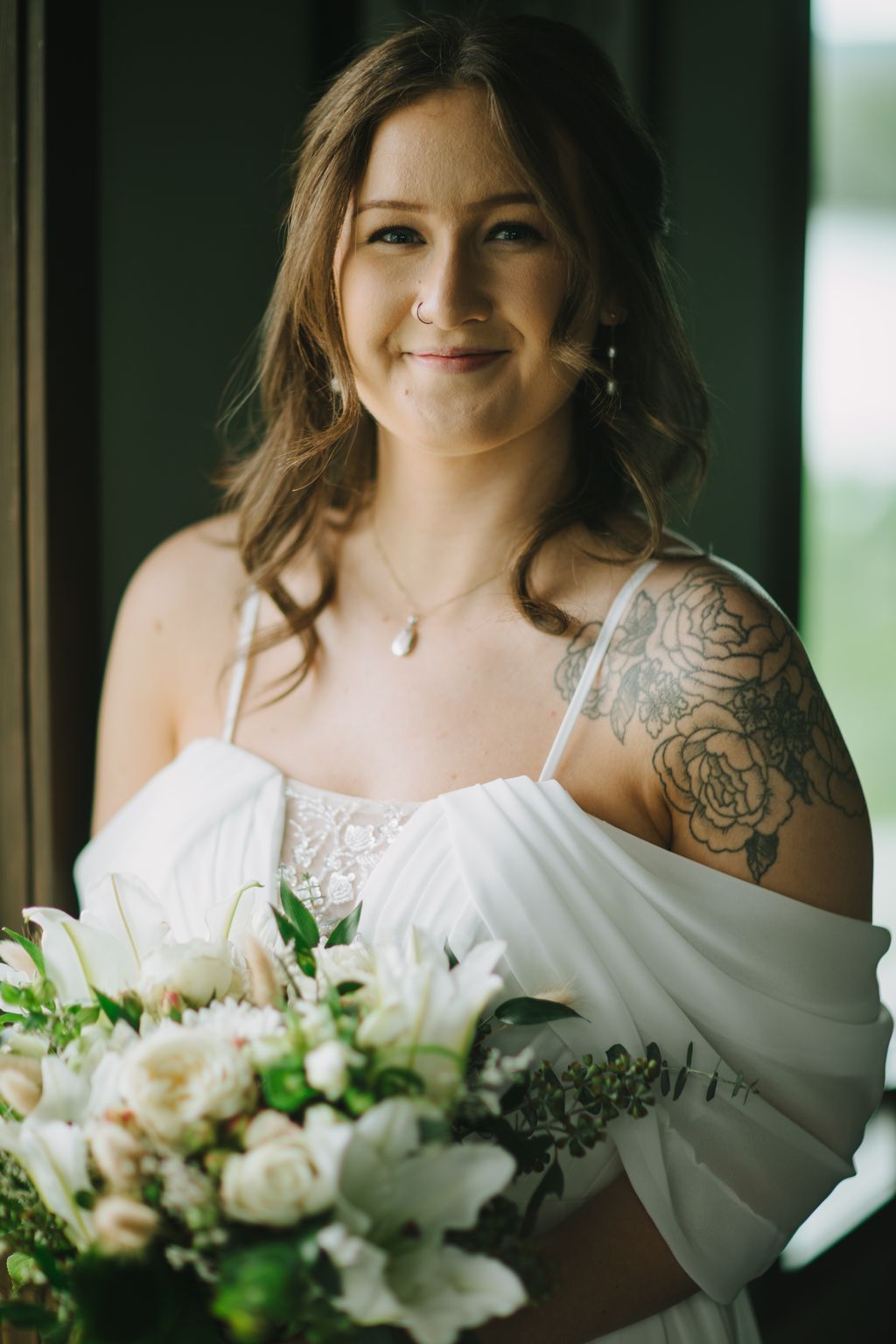 Bride in white lace dress holding white floral bouquet, smiling warmly at camera with natural window light