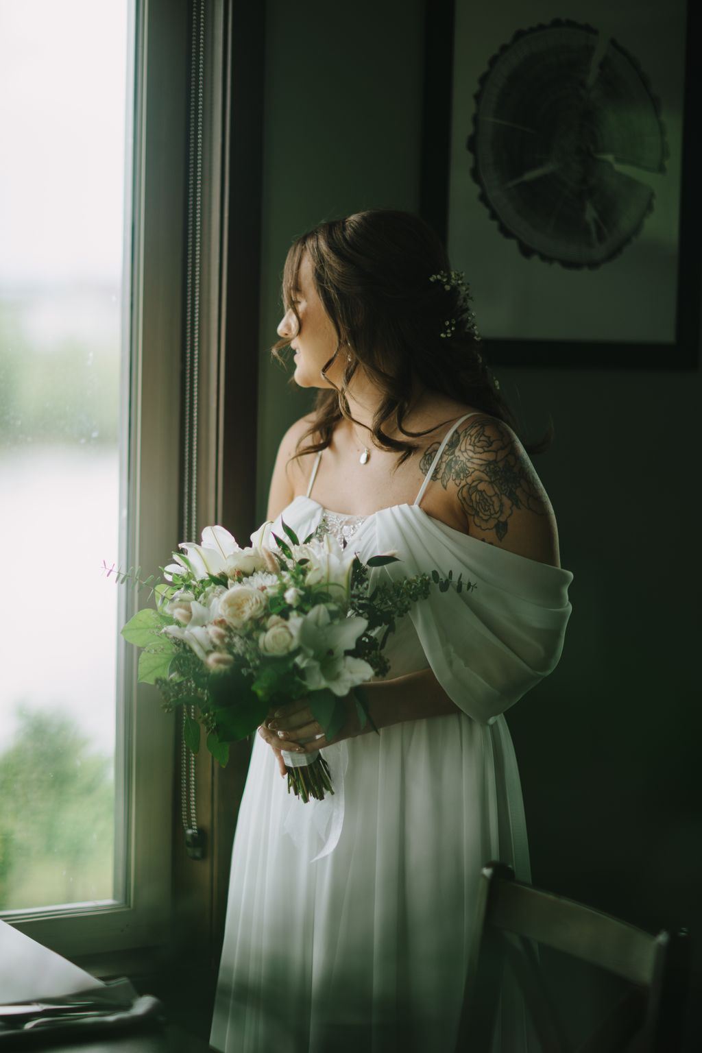 Bride in flowing white gown holding white bouquet, gazing out window with soft natural light and sage green wall backdrop