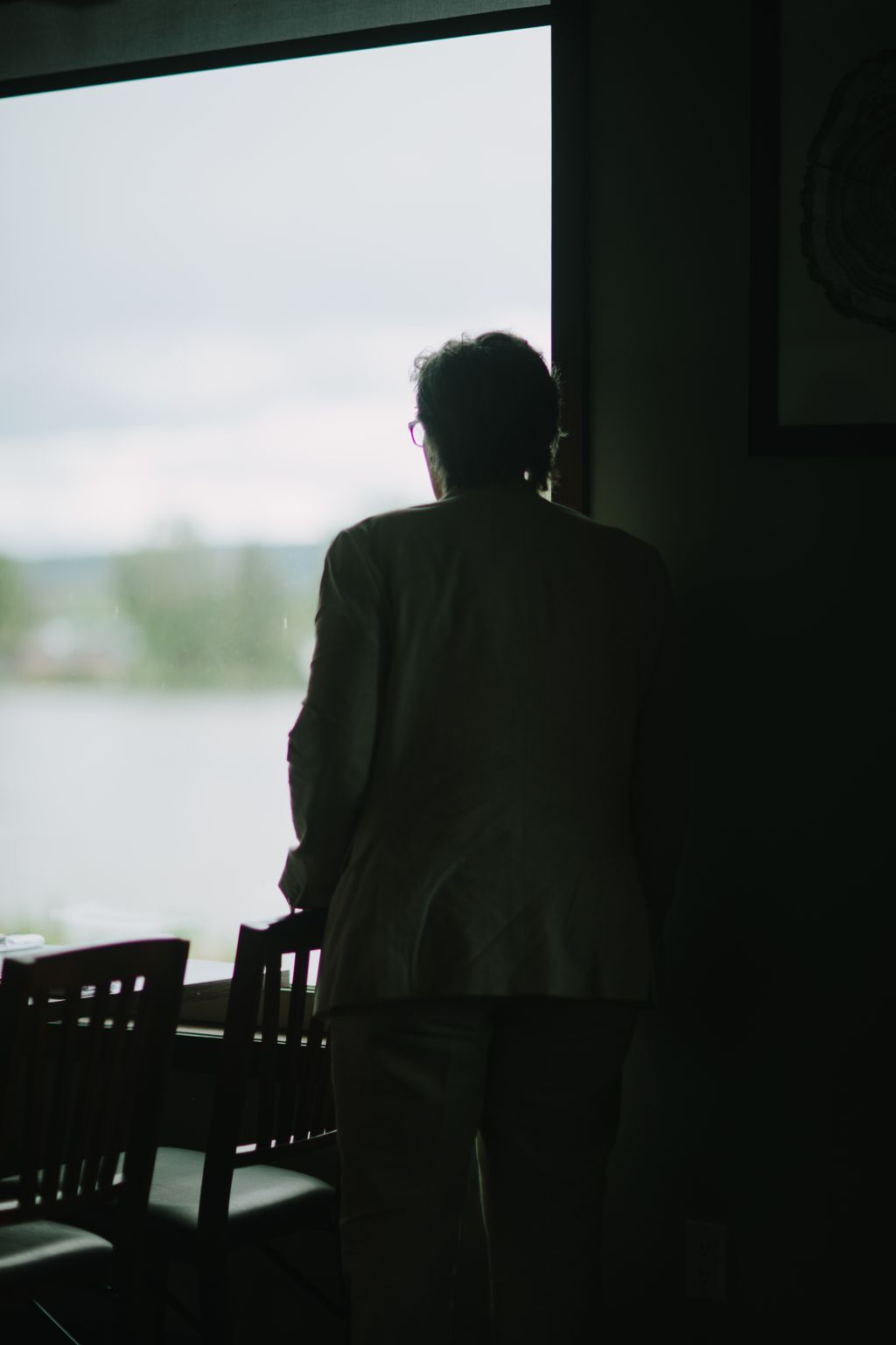 Silhouetted figure gazing out a large window toward overcast water, wearing glasses and dark clothing, intimate contemplative moment