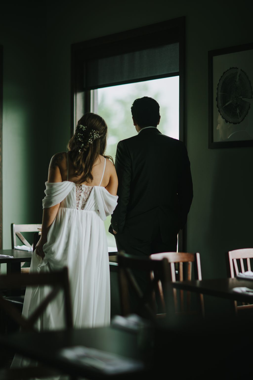 Bride and groom stand together gazing out a window with soft natural light, bride in white dress with floral hair detail, groom in dark suit