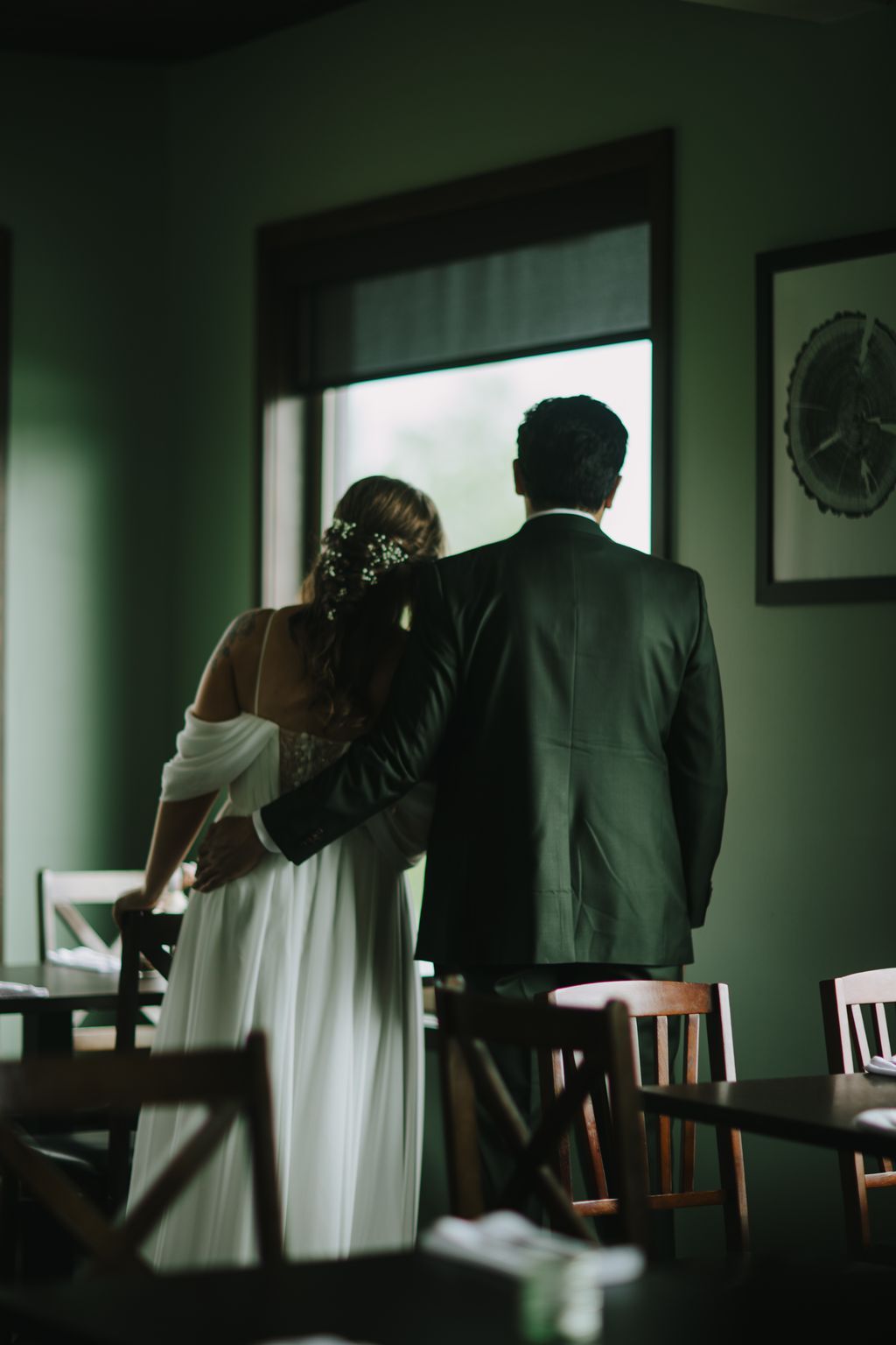 Bride and groom stand together at a window, backlit by soft natural light, viewing the landscape beyond
