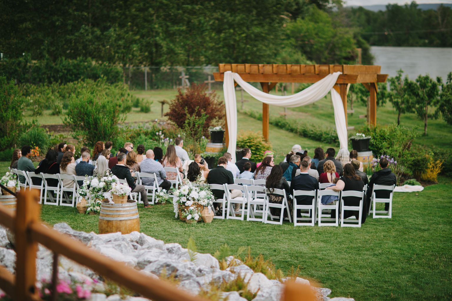 Outdoor garden wedding ceremony with guests seated under wooden pergola with white fabric draping, white flowers in foreground, lush green trees behind