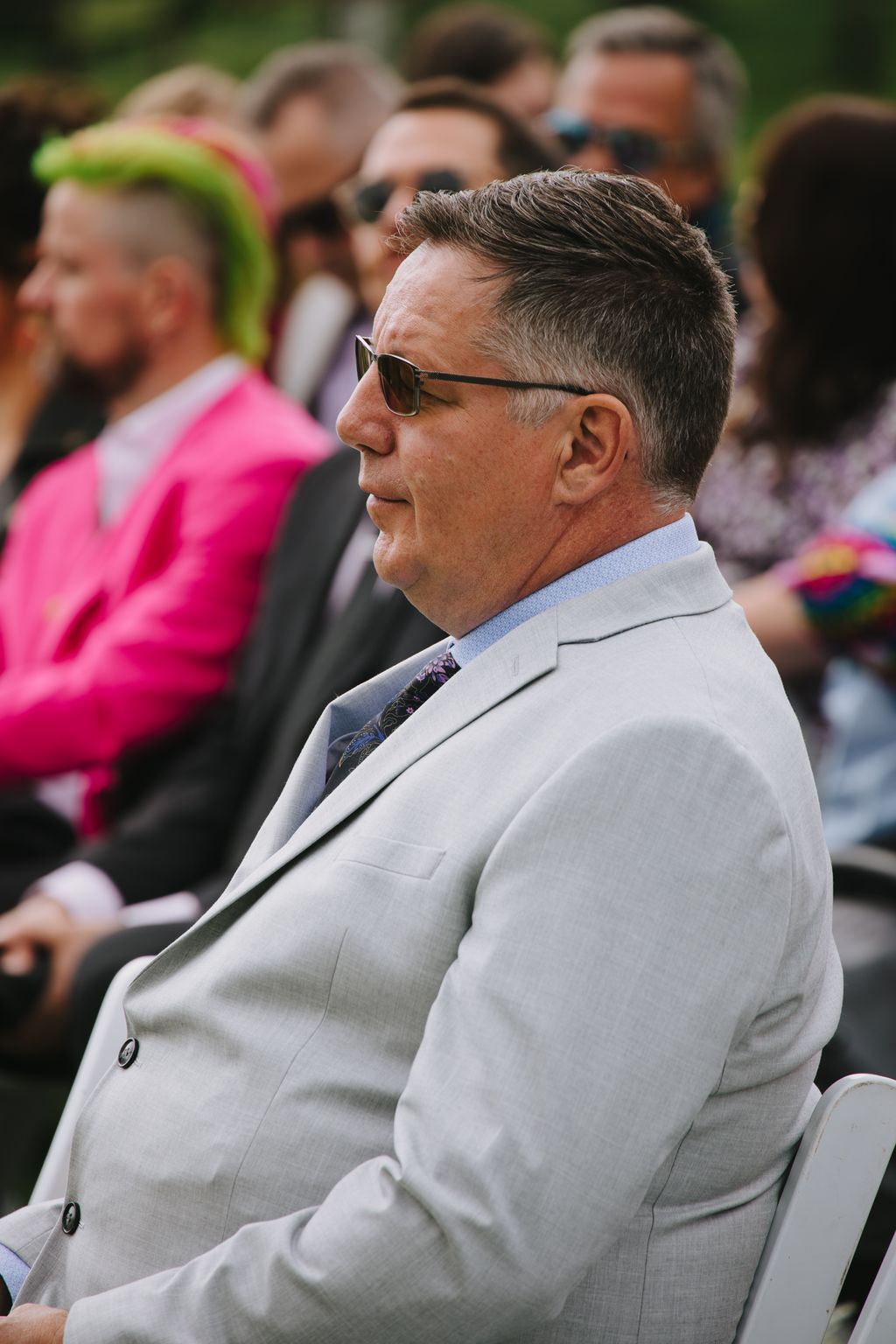 Groom in light gray suit and sunglasses smiles during outdoor ceremony with blurred guests behind him