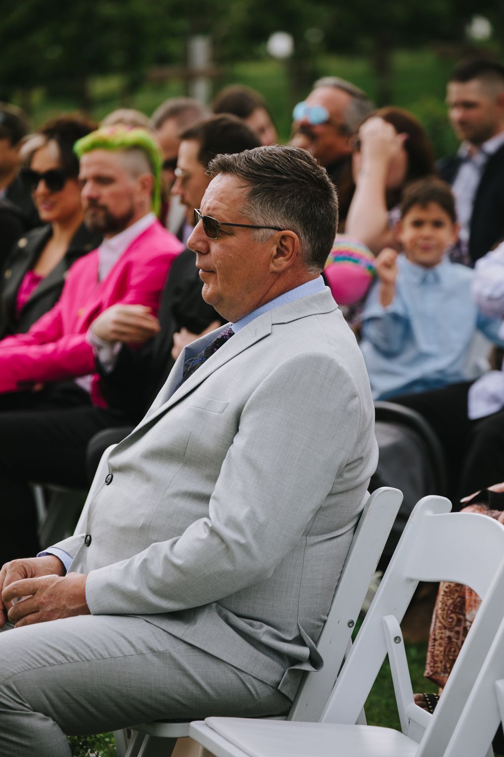 Groom in light gray suit and sunglasses seated outdoors at wedding ceremony, guests blurred in background