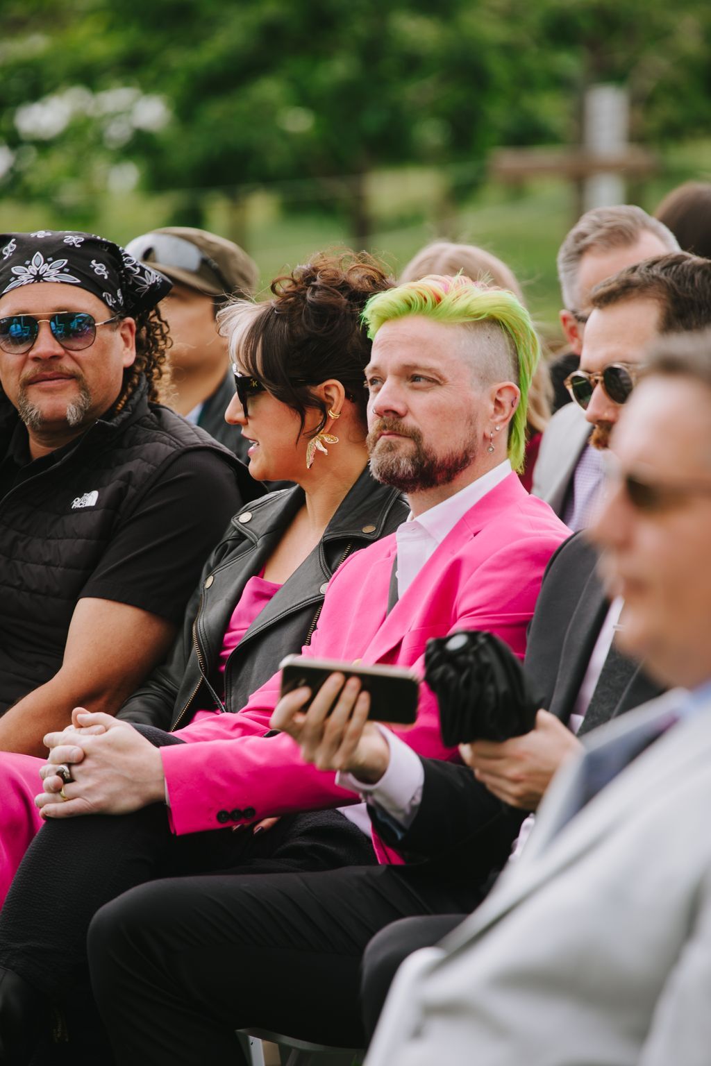 Couple in vibrant pink and black attire holding a phone outdoors, with lush green garden backdrop and natural daylight