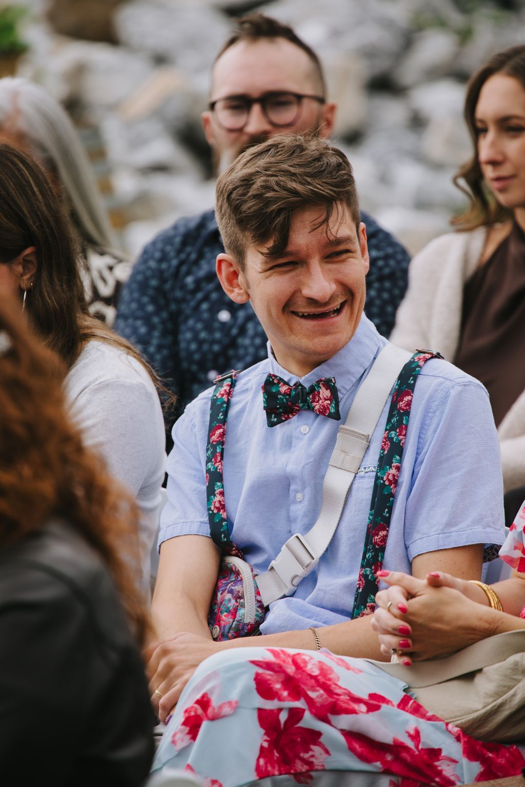 Groom in light blue shirt and floral bow tie smiles while holding bride''s hand at outdoor celebration