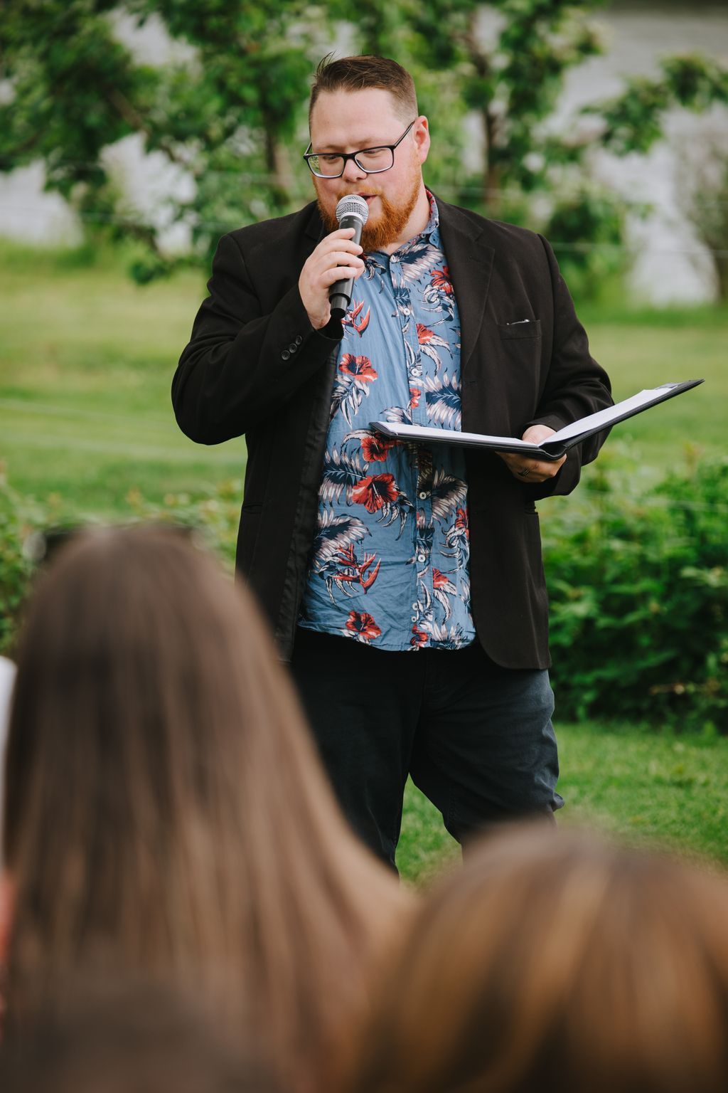 Man with red beard and glasses speaking into microphone at outdoor garden wedding ceremony