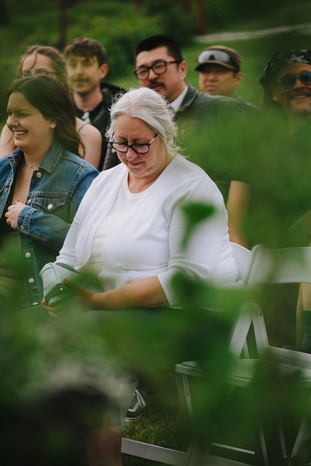 Woman in white sweater and glasses smiles warmly outdoors with guests blurred in background at garden ceremony