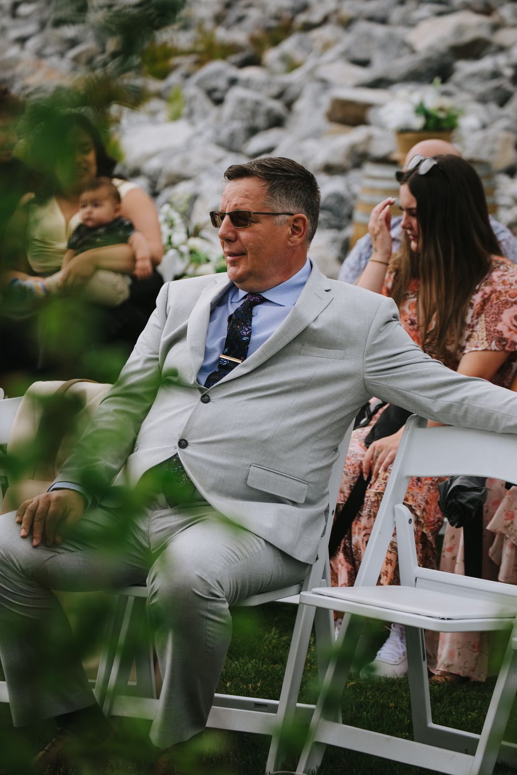 Groom in light gray suit and sunglasses seated outdoors against stone wall backdrop during wedding ceremony