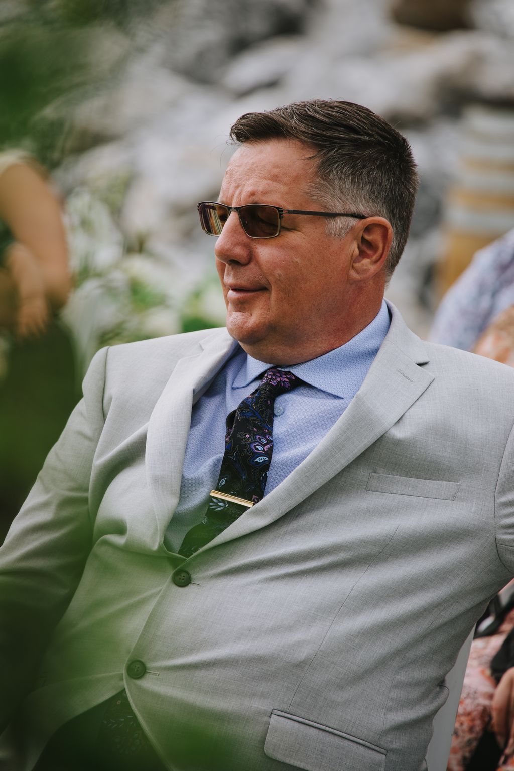 Groom in light gray suit and sunglasses with patterned tie, smiling in profile outdoors