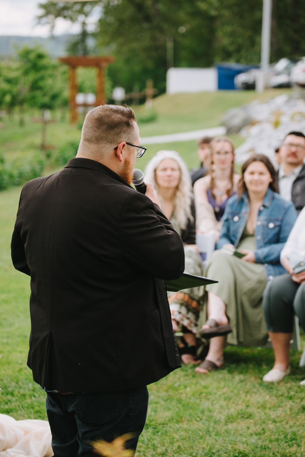 Man in black jacket speaking at outdoor ceremony with seated guests and green lawn in background