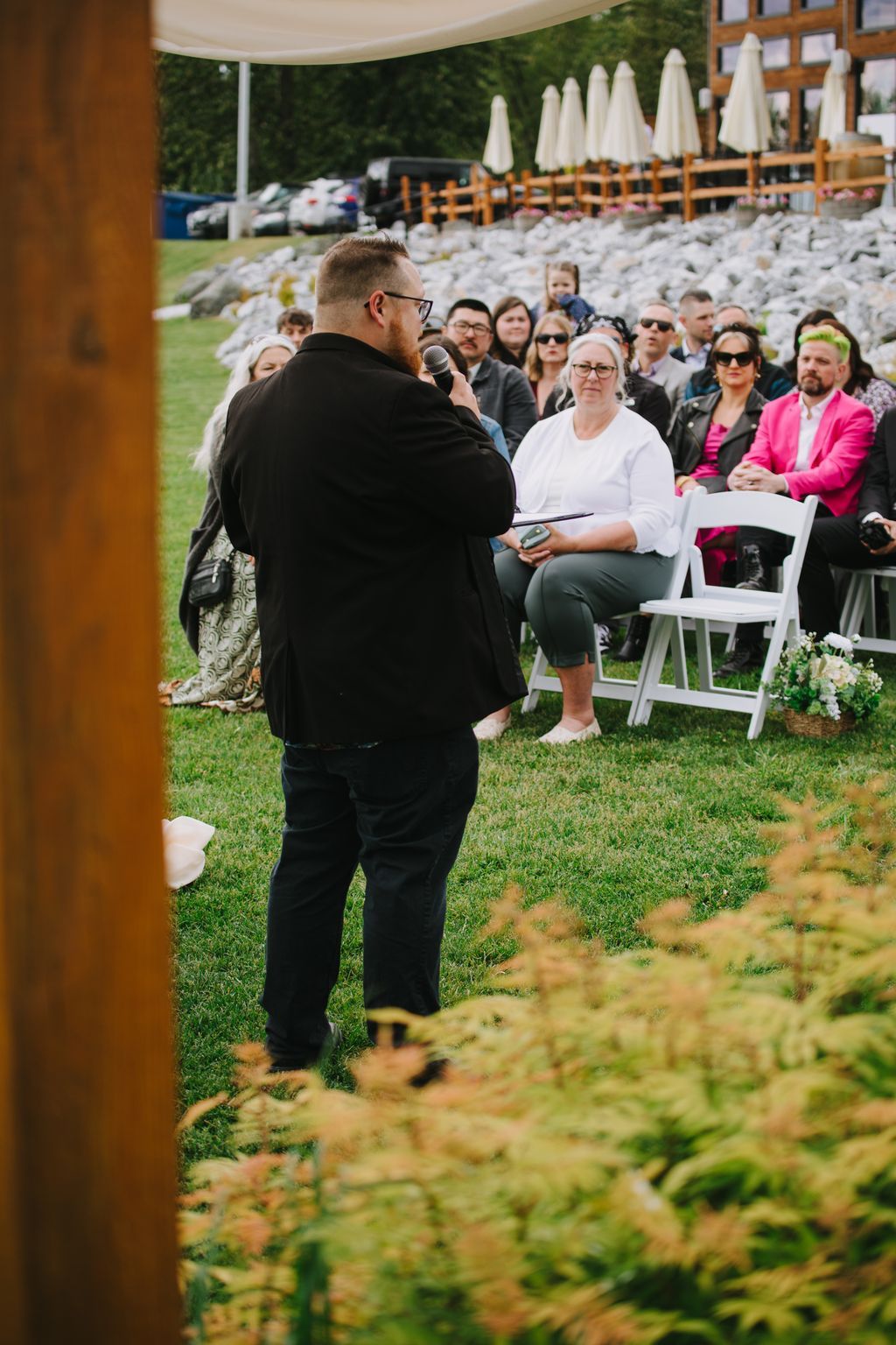 Groom speaks at outdoor ceremony while seated guests listen under white tent canopy with white umbrellas in background