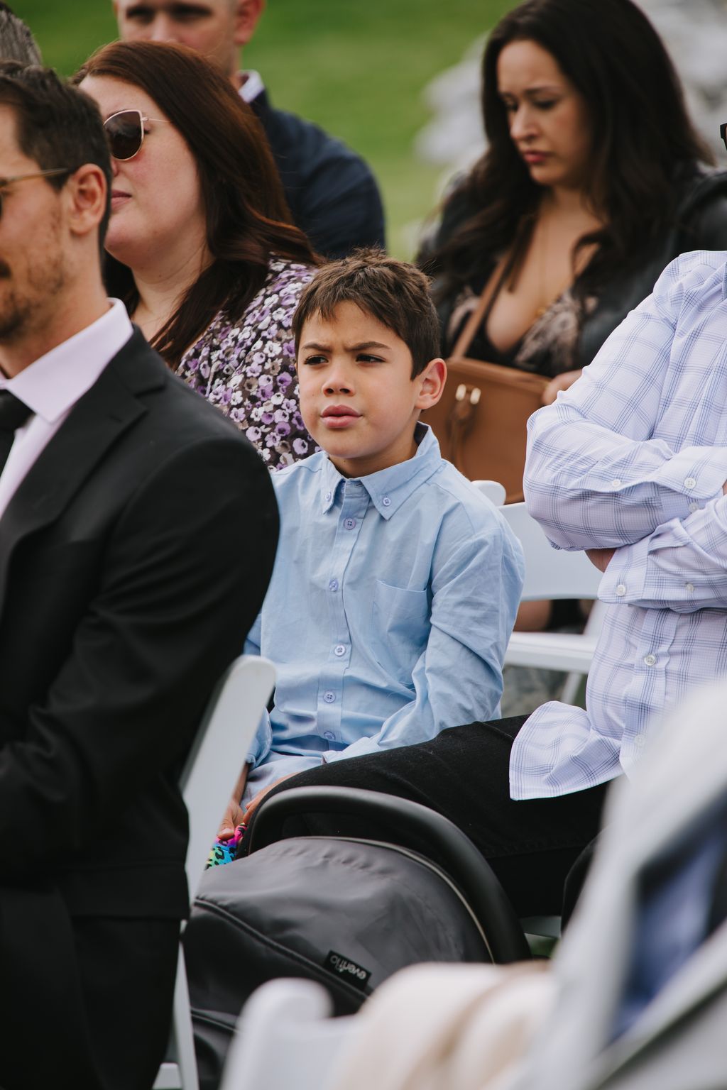 Young boy in light blue button-up shirt sitting outdoors during wedding ceremony with blurred guests and green grass in background