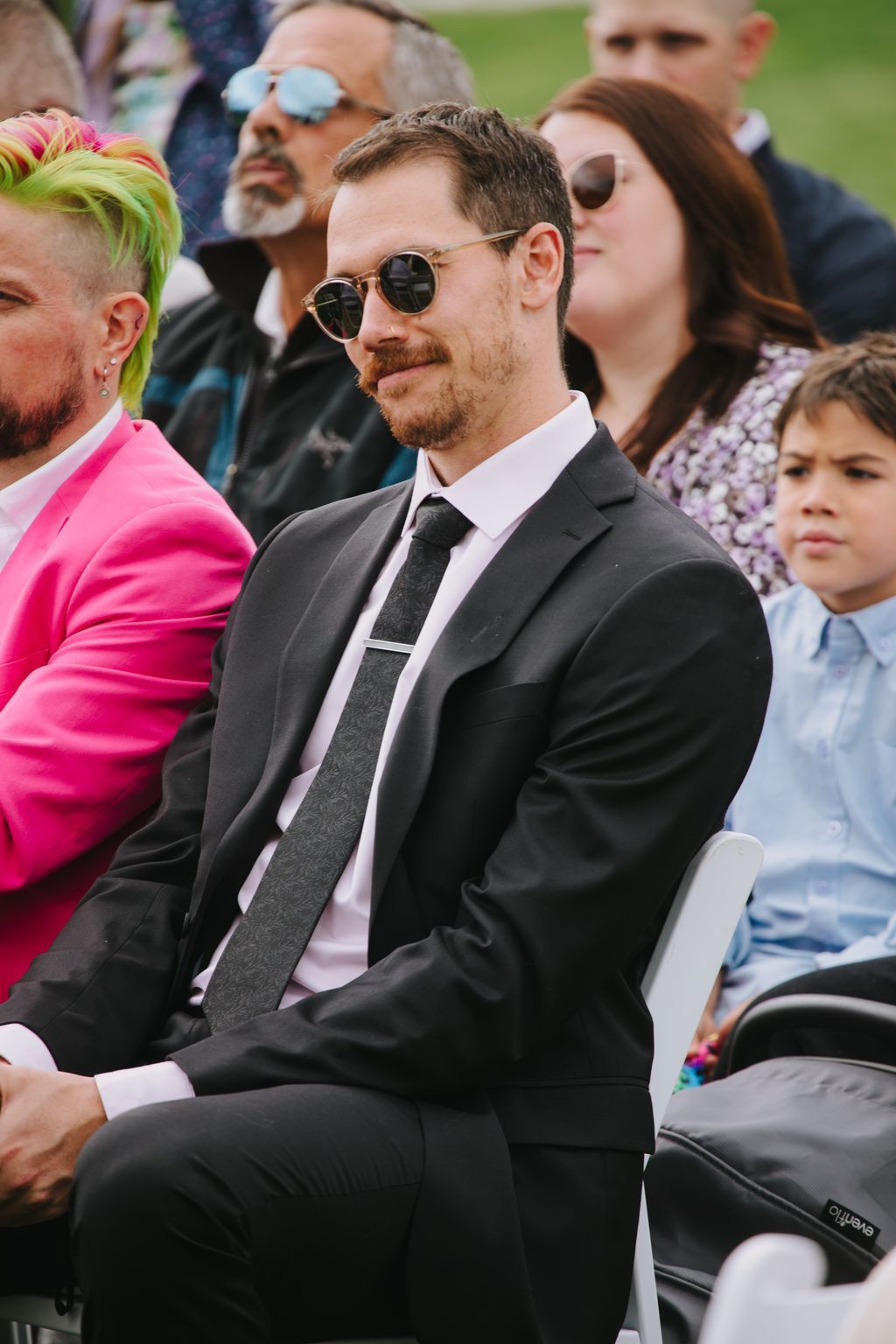 Groom in dark suit and sunglasses seated outdoors with guests blurred in background
