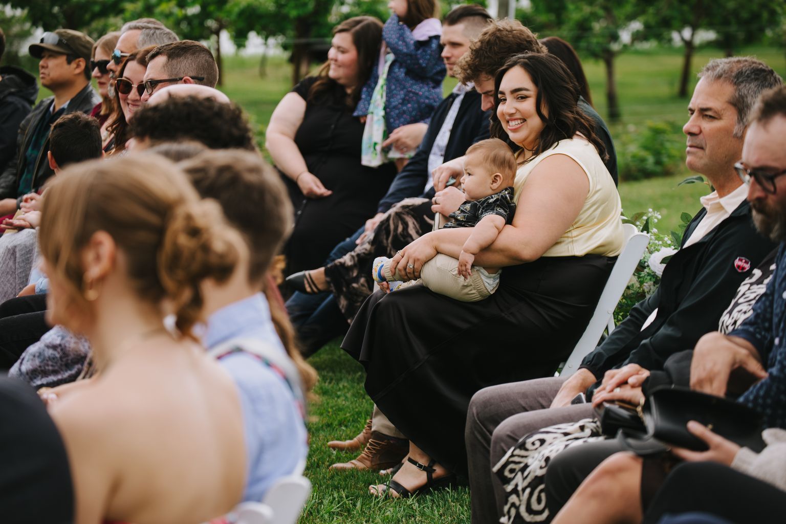 Smiling woman holding a toddler surrounded by family on a green lawn during an outdoor celebration