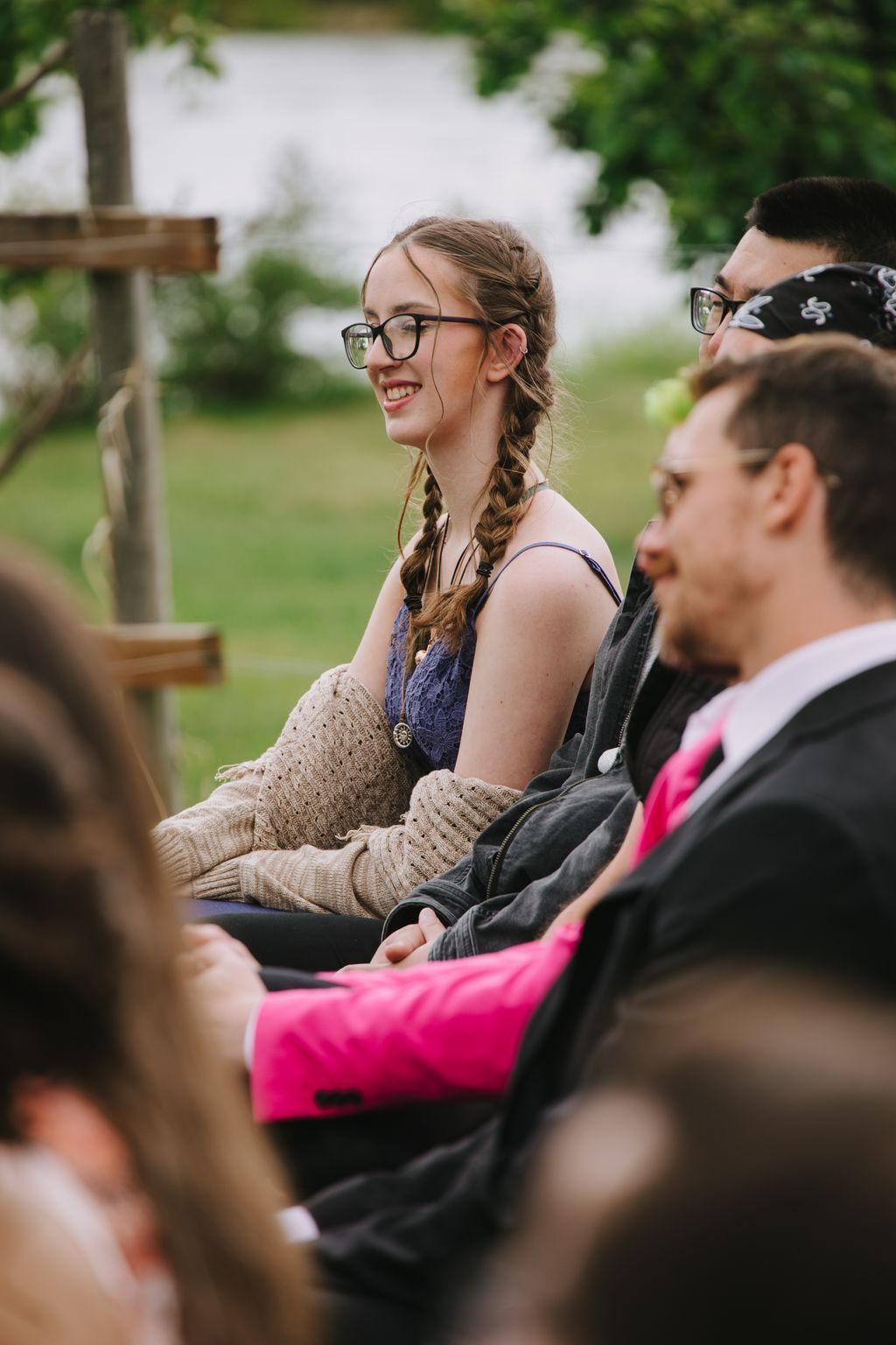 Young woman with braided hair and glasses smiles while seated outdoors with friends on a sunny day