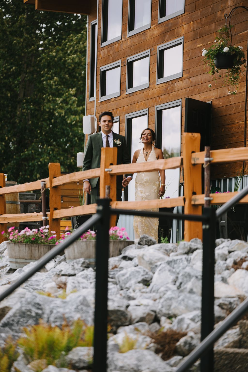 Bride and groom holding hands on wooden bridge with modern brick building and garden behind them in golden afternoon light