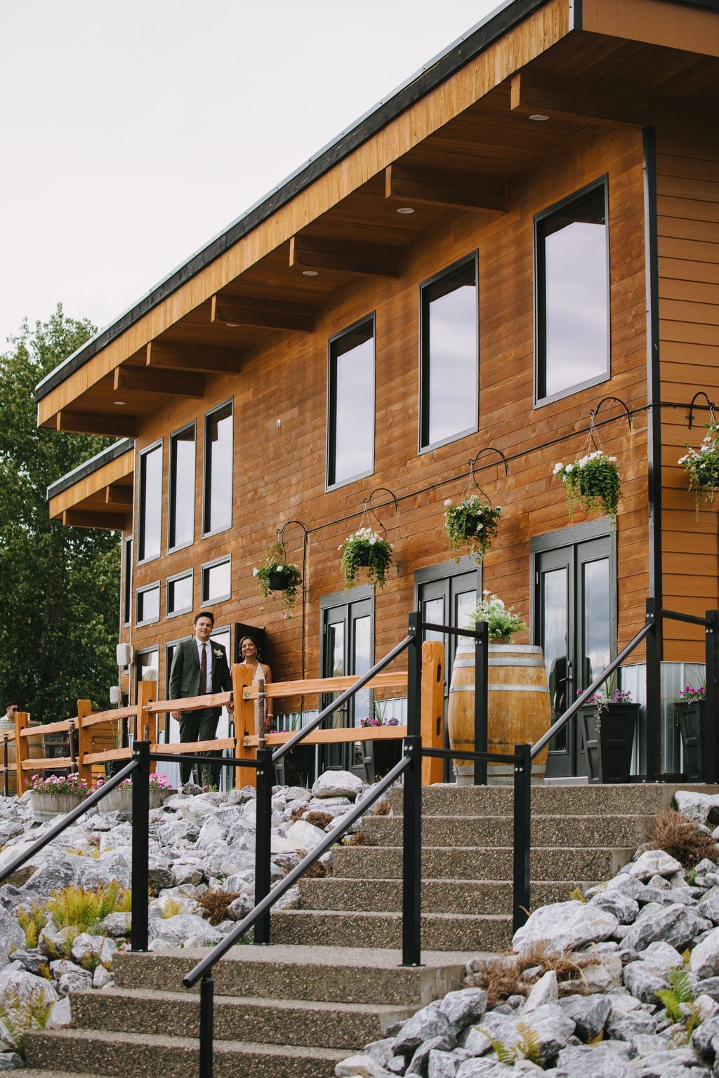 Couple in formal wedding attire standing on wooden deck of modern timber building with hanging flowers and stone steps