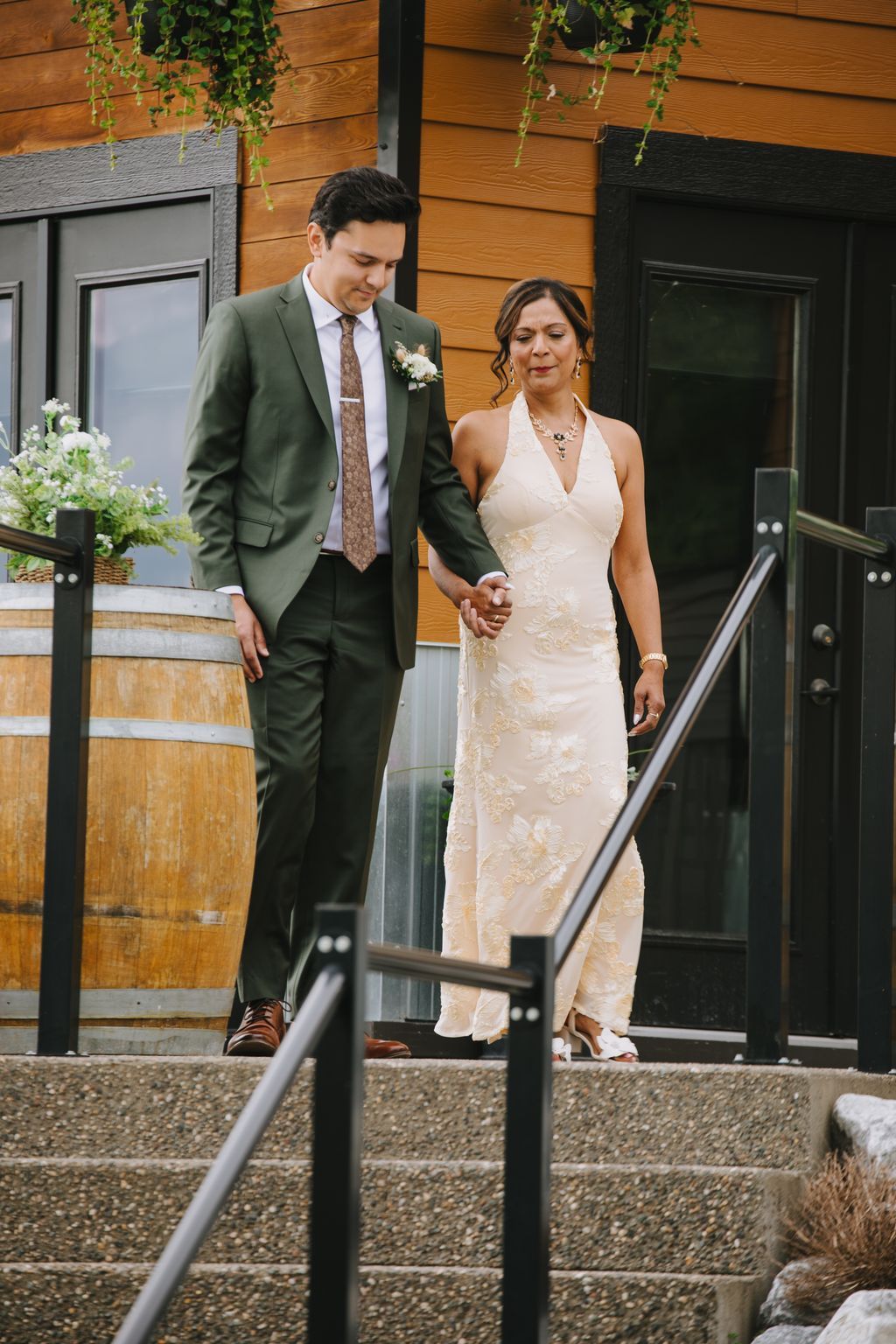 Bride in ivory halter gown and groom in sage green suit hold hands descending wooden steps at their wedding ceremony