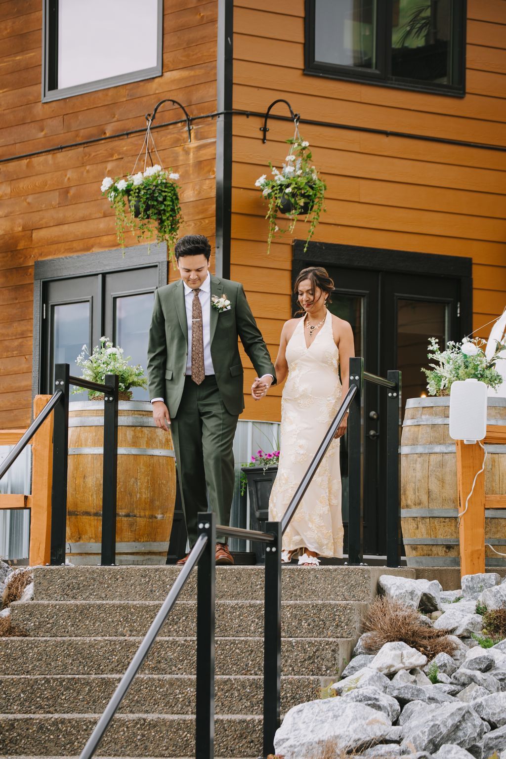 Bride in ivory lace gown and groom in sage green suit descend wooden stairs at modern barn venue, hanging floral baskets above