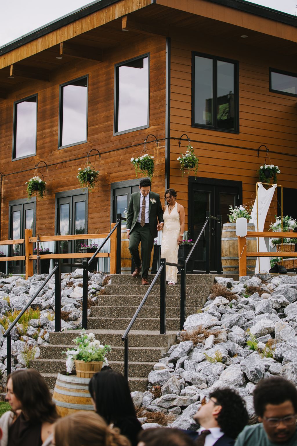 Bride and groom descend wooden lodge steps hand in hand, surrounded by hanging flowers and stone landscaping