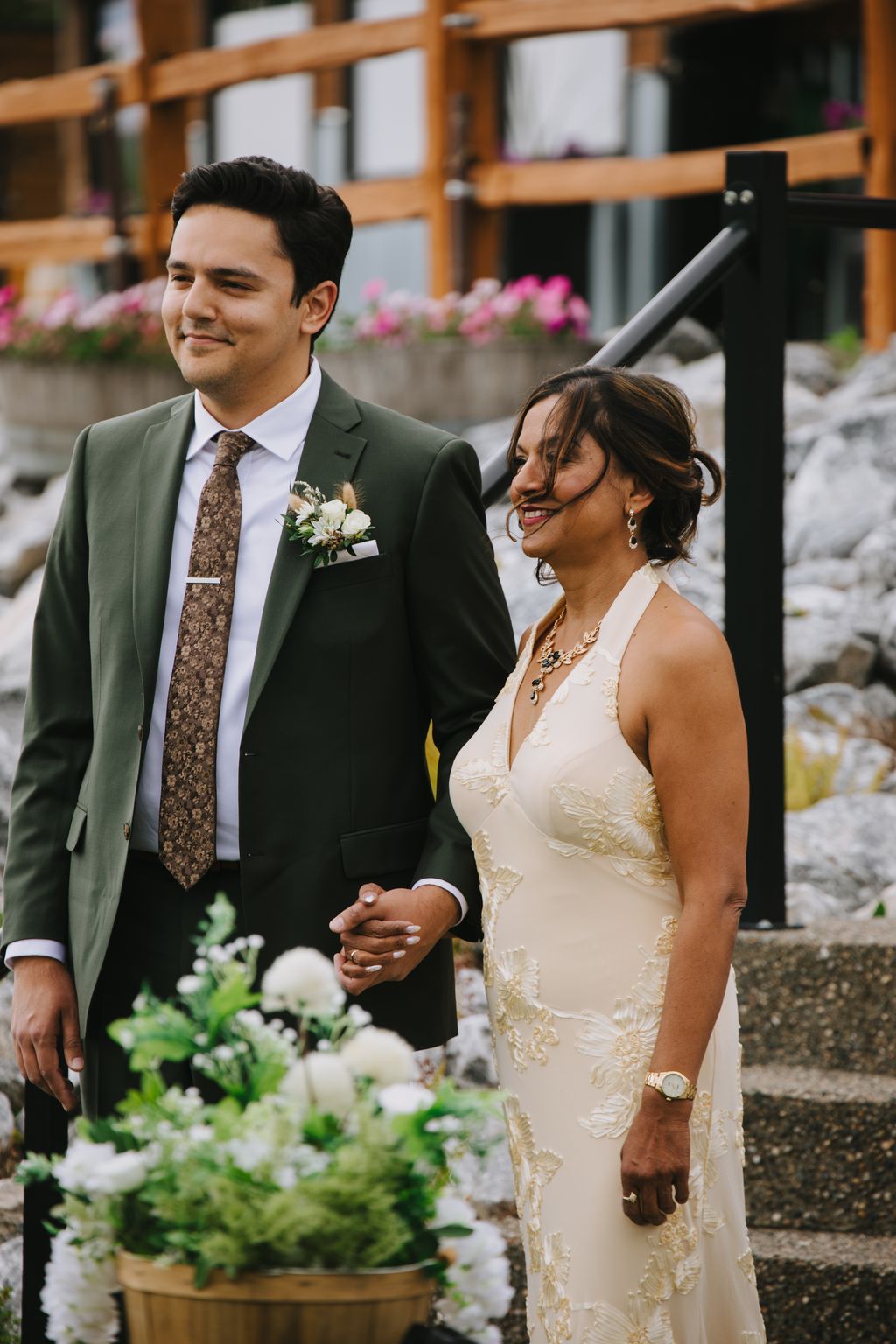 Bride in beaded ivory gown and groom in sage green suit hold hands at outdoor ceremony with white florals and wooden structure backdrop